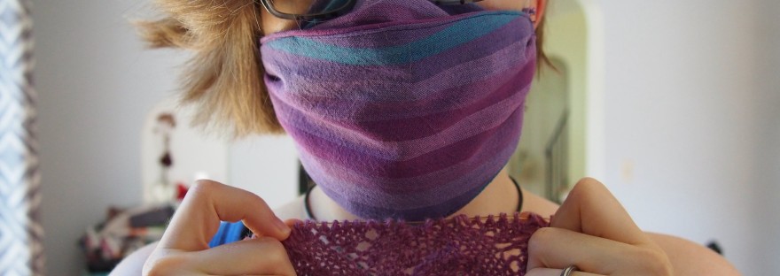 Woman wearing glasses and a striped cloth mask, holding a lacy knitting project that is still on the needles and looking pensive.